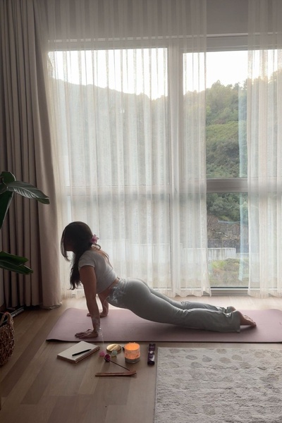 A woman performs a cobra stretch on a yoga mat in a sun-drenched room with incense and a journal, emphasizing a peaceful wellness lifestyle.